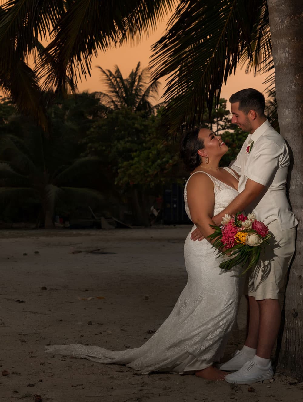 A couple shares a joyful moment during their beach wedding at sunset, surrounded by tropical foliage.
