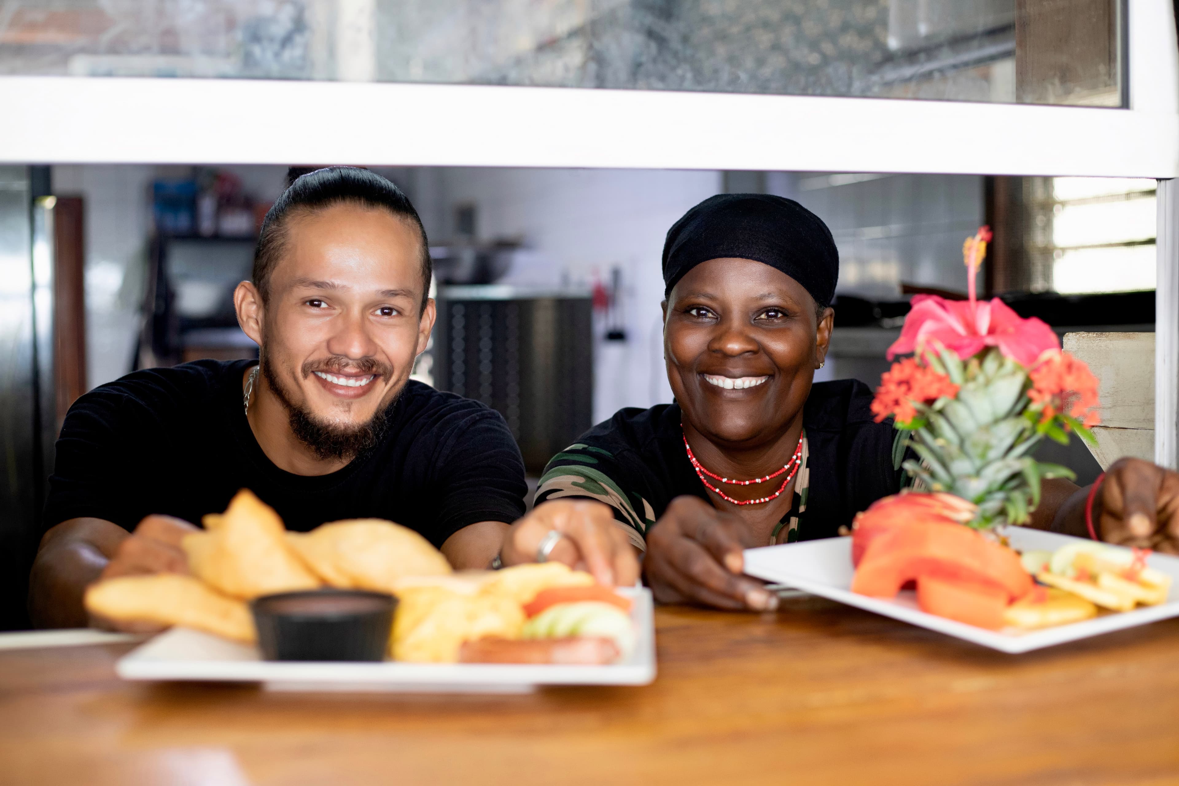 Two smiling individuals present trays of food in a cozy eatery setting.