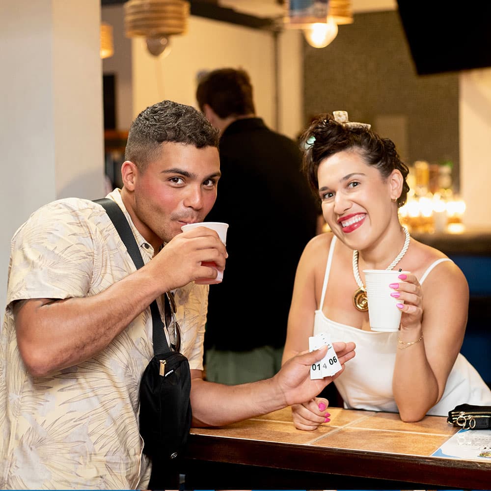 A man and woman smile while holding cups at a bar, with the woman displaying a small ticket.