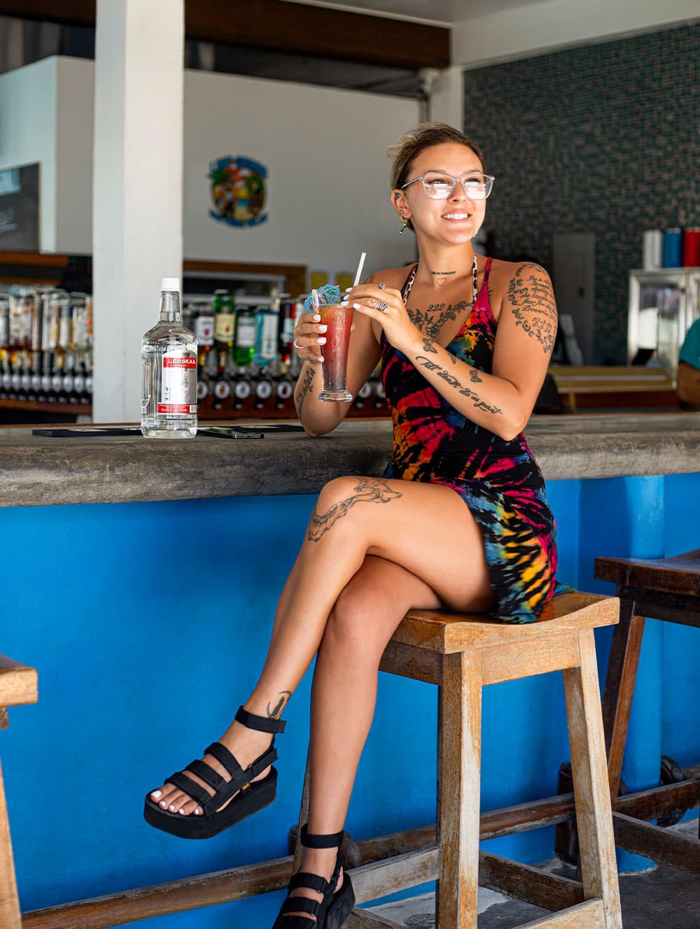 A woman with tattoos sits on a bar stool, holding a colorful drink and smiling at the camera.