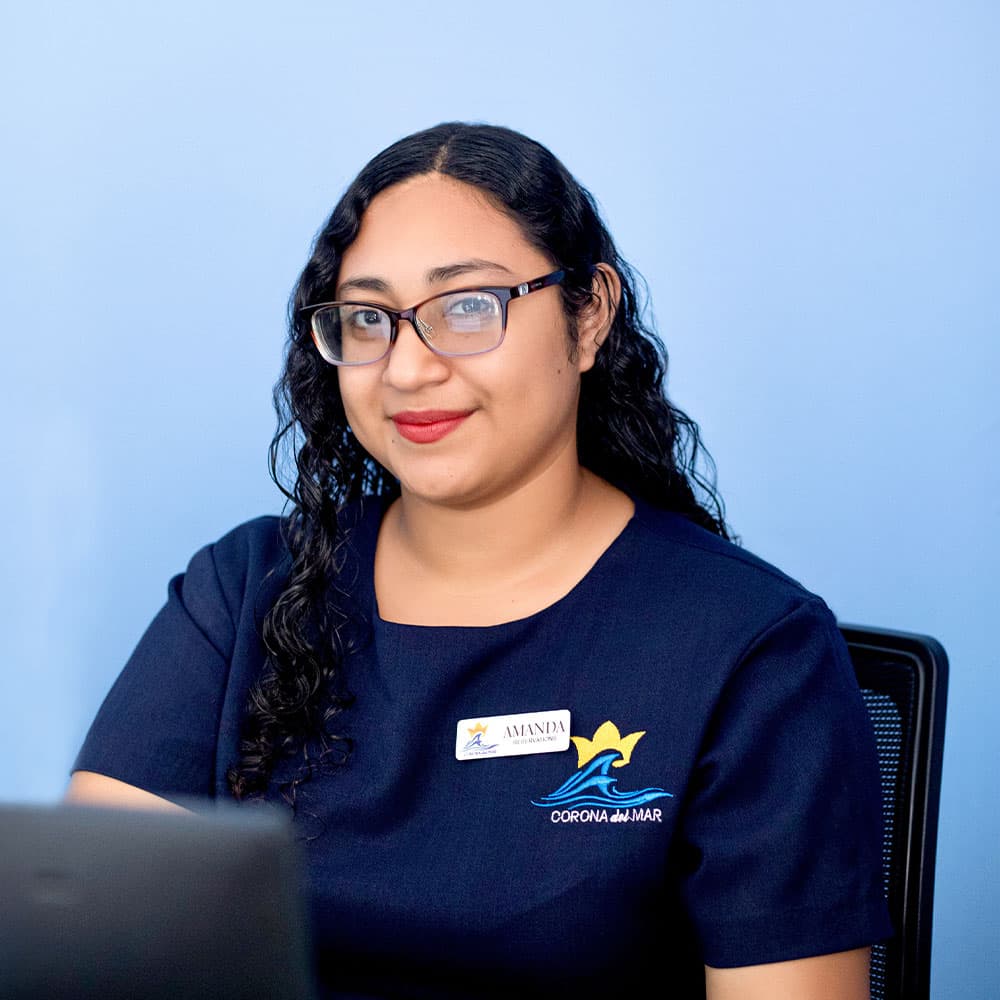 A woman with curly hair and glasses smiles while seated at a desk, wearing a dark uniform with a logo.