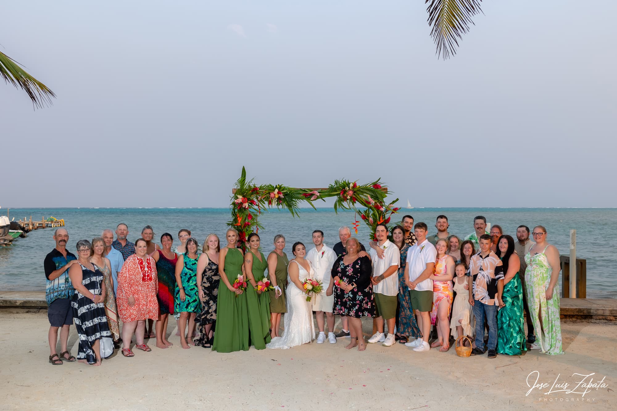 A large group of people gathered at a beach wedding, standing in front of a floral arch with the ocean in the background.