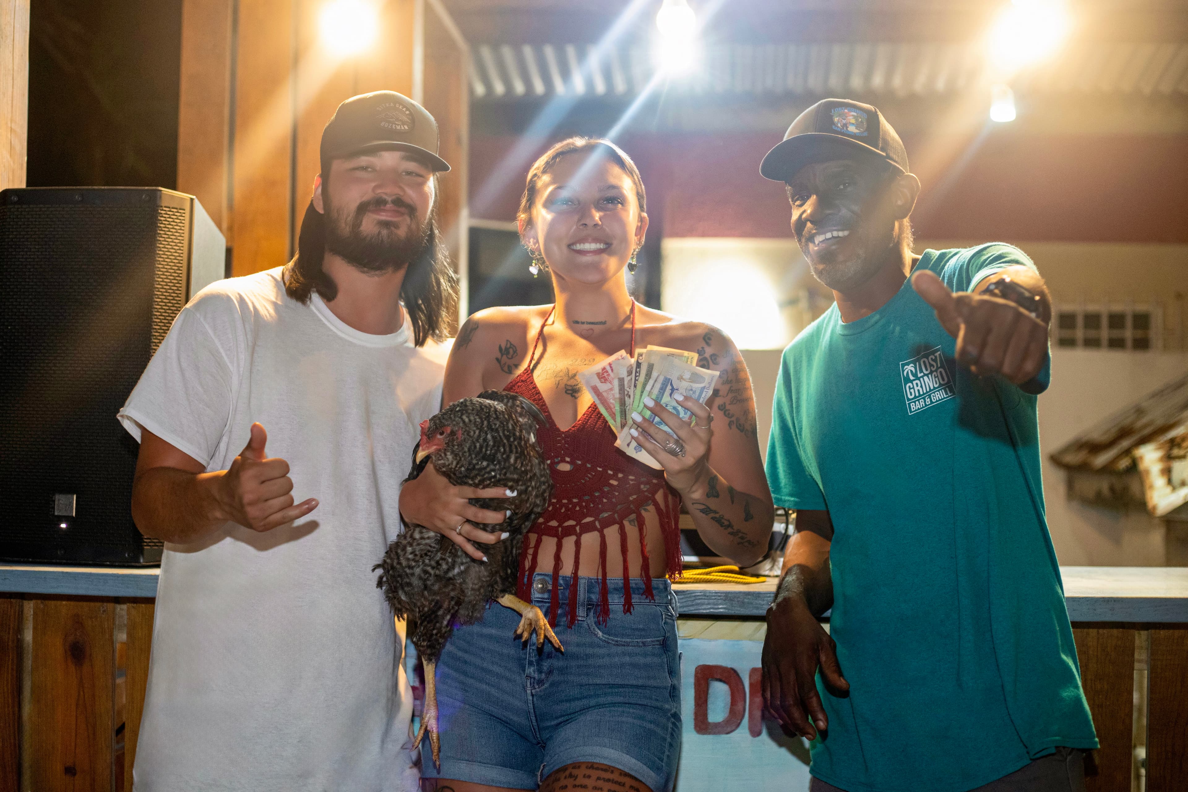 Three people smile together, one holding a chicken and another displaying cash, in a lively setting.