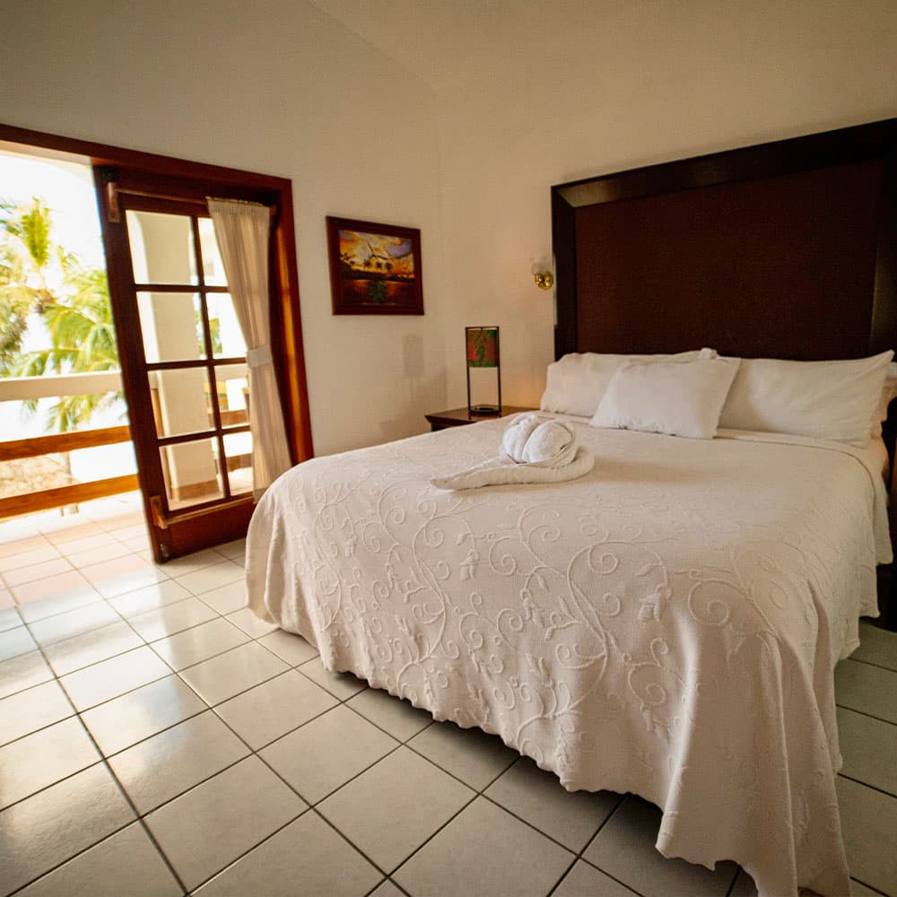 A bright hotel room featuring a white bedspread, pillows, and a balcony view.