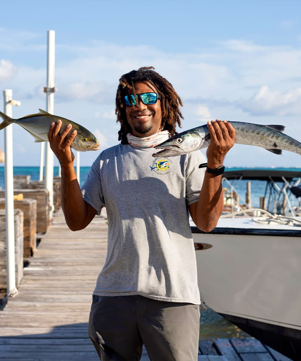 A man smiles on a dock, holding two freshly caught fish.
