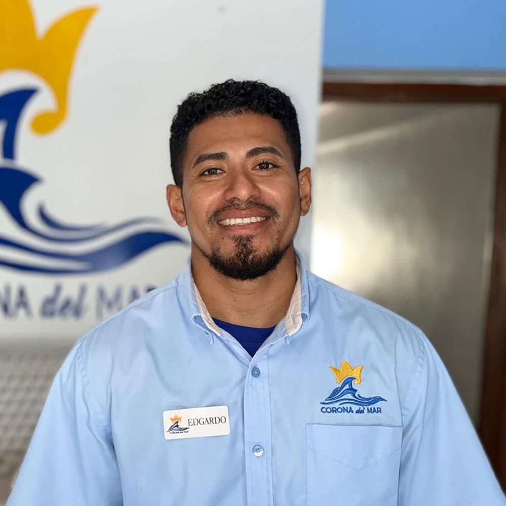 A smiling man in a blue shirt with a name tag stands in front of a logo for "Corona del Mar."