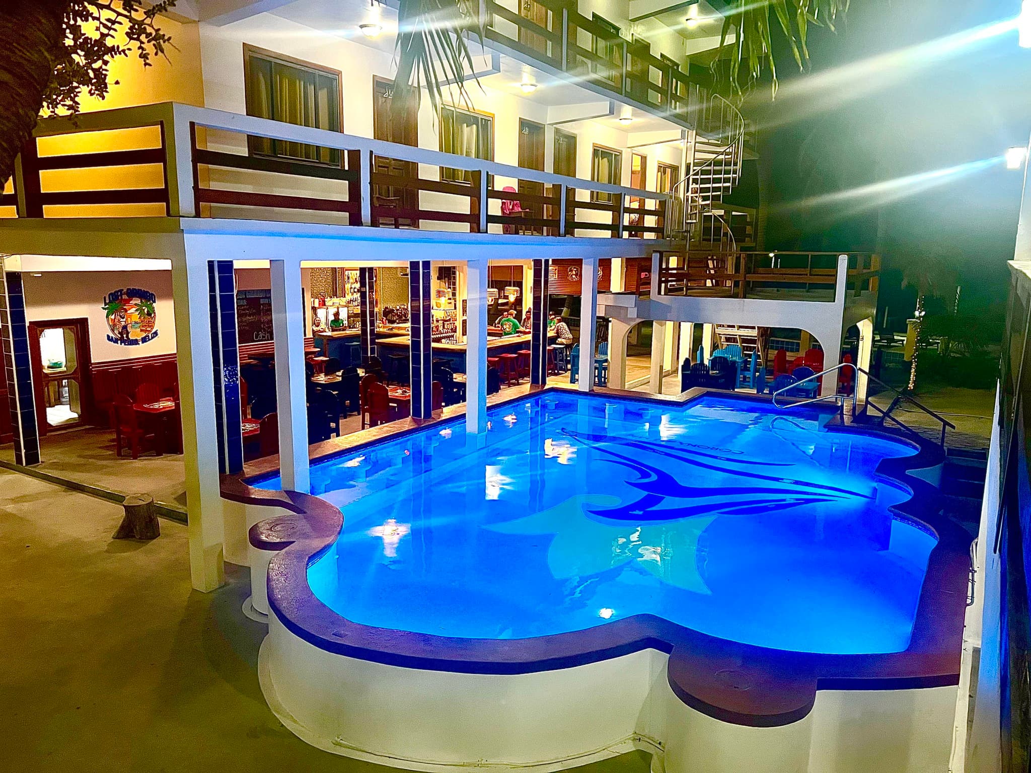 A well-lit pool area surrounded by a two-story building with outdoor seating and a spiral staircase.