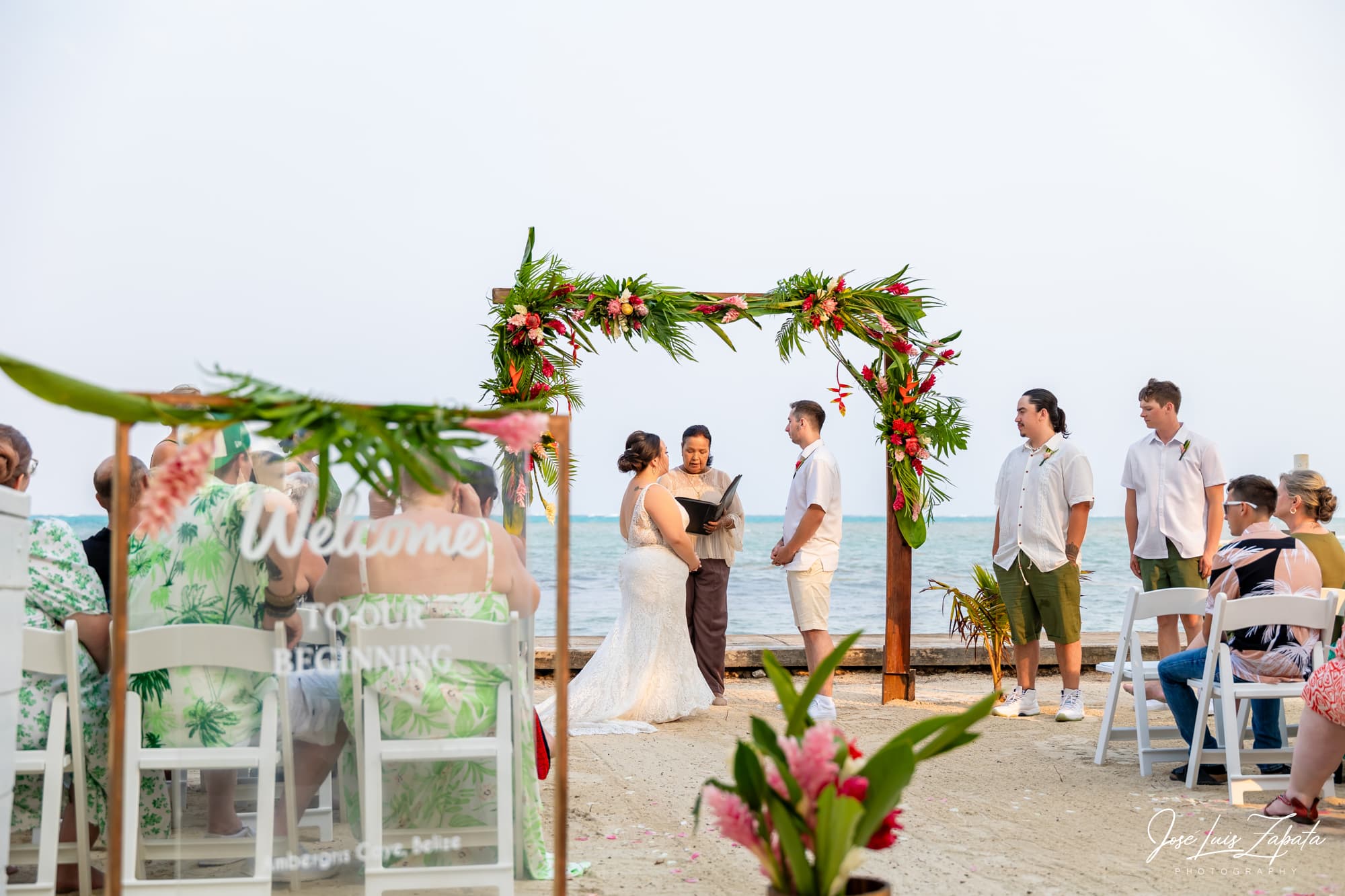A couple stands under a floral arch at a beach wedding while guests look on.