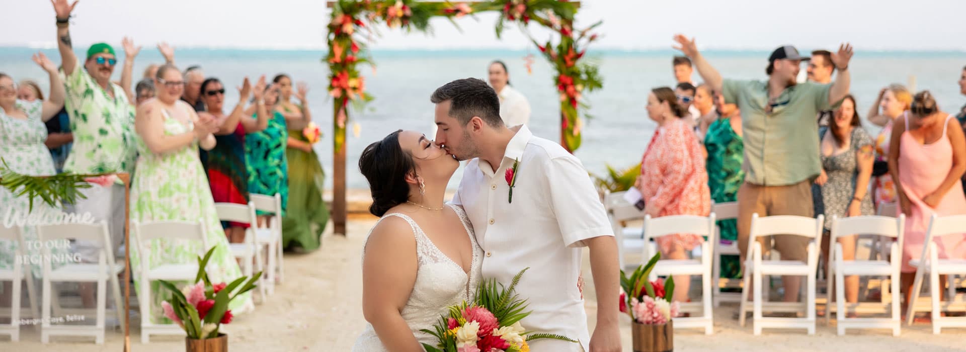 A bride and groom kiss at their beach wedding as guests cheer in the background.