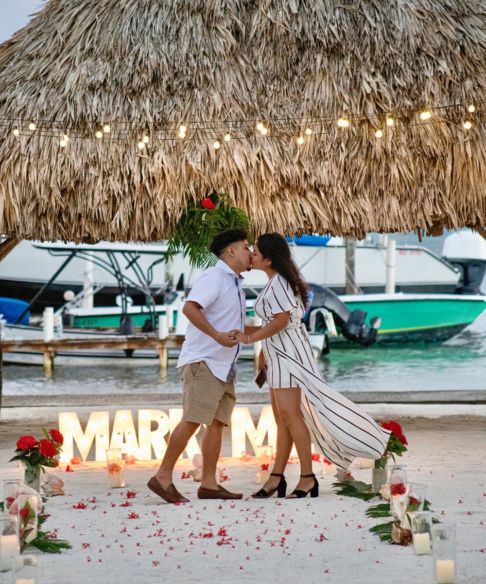 A couple kisses under a thatched roof, surrounded by candles and flowers on a beach.