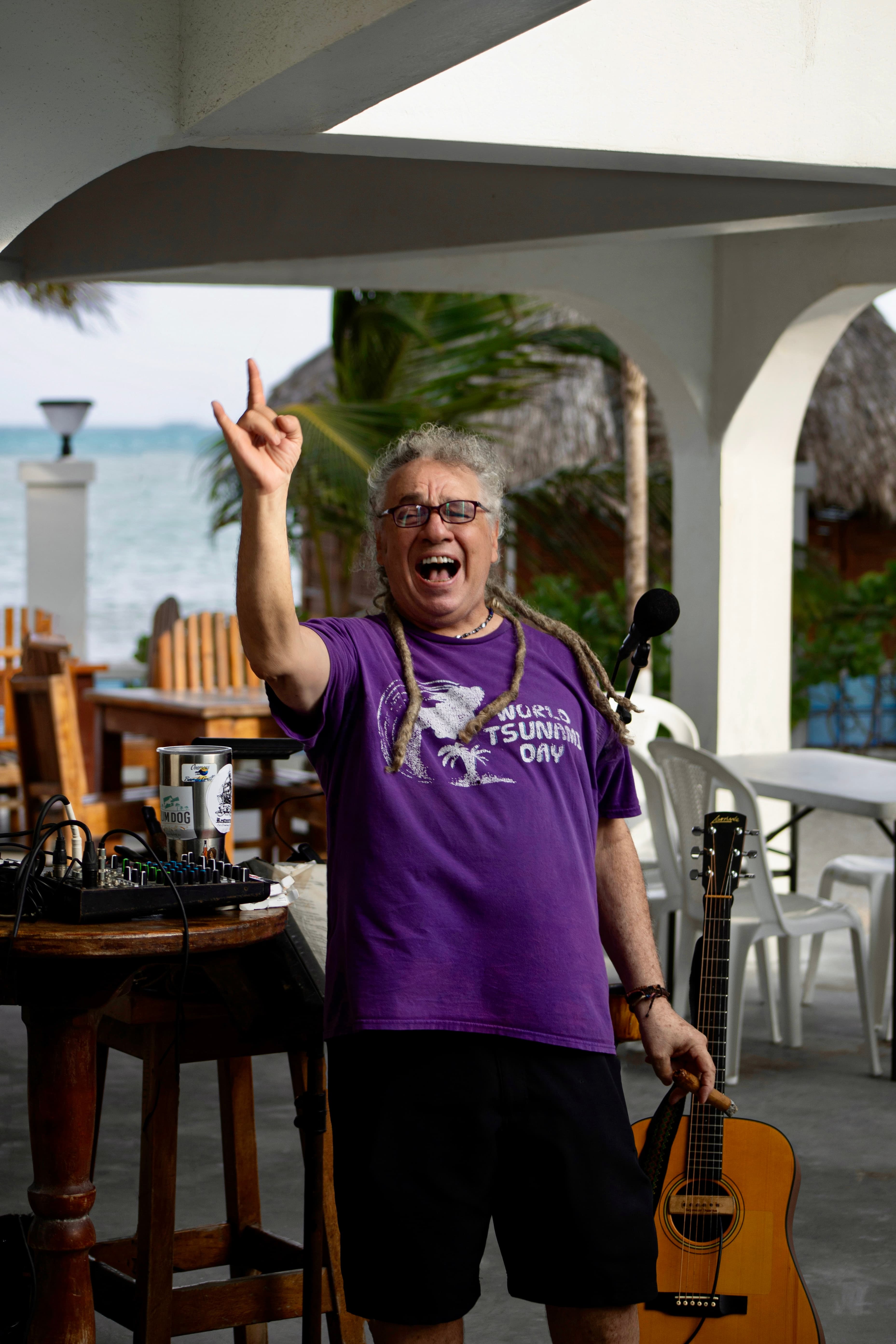 A smiling man with long hair and glasses holds a guitar while gesturing enthusiastically in a beachside setting.